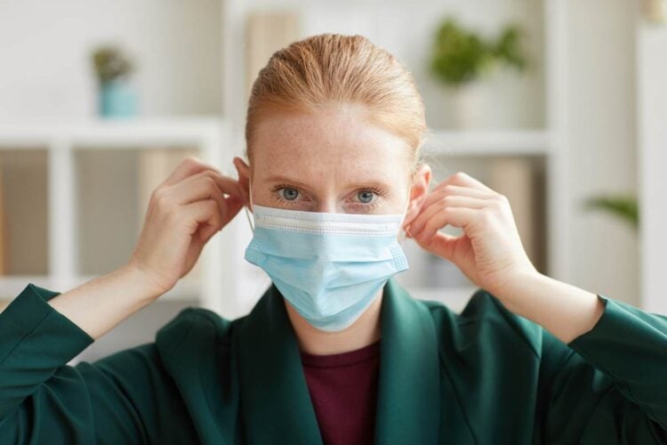 Head ad shoulders portrait of young businesswoman putting on face mask and looking at camera while working in post pandemic office
